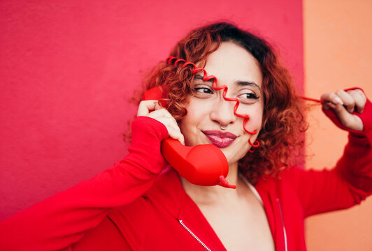 Redhead with curly hair playfully talking on red telephone indoors