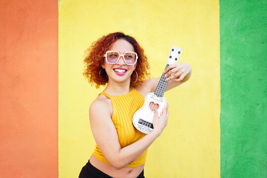 Smiling redhead with ukulele in summer fashion against colorful wall