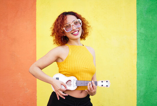 Smiling redhead with glasses playing ukulele in front of colorful wall