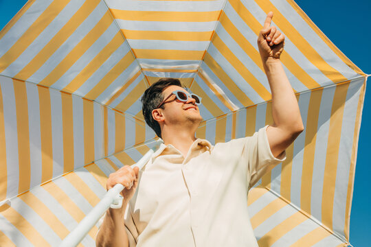 Smiling man in sunglasses under striped umbrella on sunny summer day