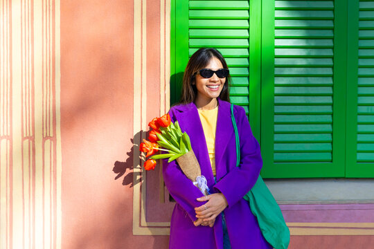 Smiling woman with tulips and sunglasses by green window outdoors