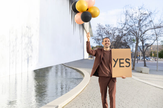 Happy man holding YES sign and balloons outdoors celebrating success