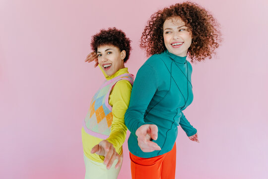 Two friends dancing and smiling back to back on pink background