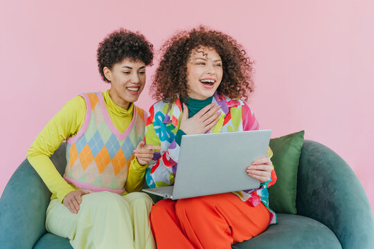 Women friends with curly hair laughing together on sofa with laptop