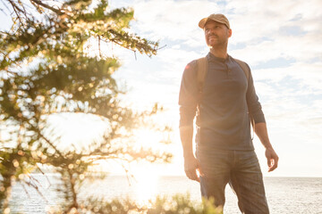 Naklejka premium Hiking, travel and walk with man in nature for trekking adventure, backpacking and seaside trail. Tree, summer vacation and sunshine with person or hiker outdoor for ocean, explore and tourism