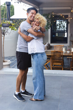 Couple embracing, standing on tiled patio, surrounded by plants, table, sofa, string lights