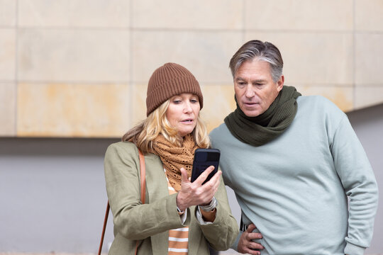 Senior couple studying smartphone while standing in front of beige tiled facade, holding bag
