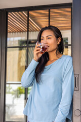 Asian adult woman standing at glass door wearing light blue top holding clear cup, slatted shadows © wavebreak3