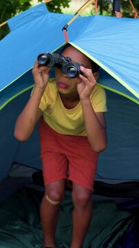 Vertical video: Spotting motion, child in tee peering with binoculars in yard tent, exiting smiling