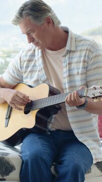 Vertical video: Placing guitar on thigh, man fretting and strumming by bright window, practicing