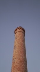 Tall stone lighthouse tower rises into a clear pastel evening sky, softly lit by warm light, emphasizing its cylindrical form and small vertical windows. © Aperezer