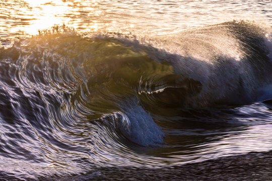 Curling ocean wave illuminated by warm golden sunlight at the shoreline, capturing motion, texture, and shimmering reflections in a tranquil coastal seascape.