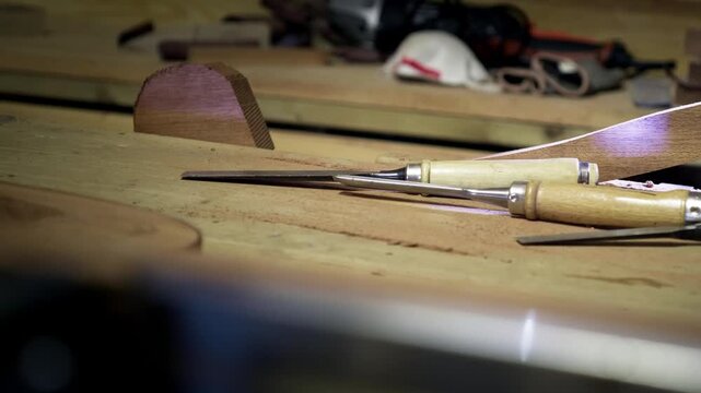Selection of sharp carving chisels resting on a workbench alongside a partially finished timber project in a carpentry studio. Still life in slow motion.