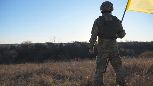 Rear view of male soldier in military uniform holds a waving national flag. Ukrainian army man stands with lifting blue-yellow banner at field. End of war in Europe. Invasion of territory Ukraine
