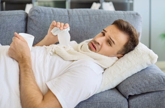 Sick man with stuffy runny nose, allergy or viral fever resting on sofa at home. Sad person in scarf holding tissue and mug, holding mug of hot drink, suffering from fatigue, headache and inflammation