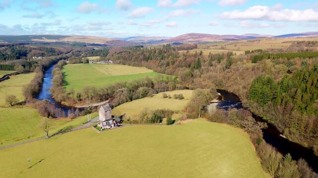 The River Esk winds through green fields and dense trees at Gilnockie Tower in the Scottish Borders. This scene shows a clear view of the landscape near Langholm on the A7. Hills rise in the backgroun