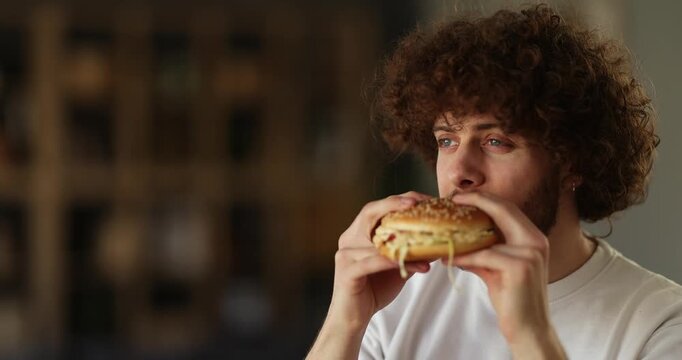 A young man enjoys a burger while eating it at home.