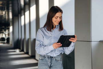 Young woman with long brown hair wearing a light blue shirt holds a tablet while standing in a modern corridor with natural light streaming through windows