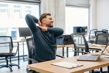 Male office worker in blue sweater stretches in modern workspace with wooden desks, laptop, and empty chairs, bright windows providing natural light © standret