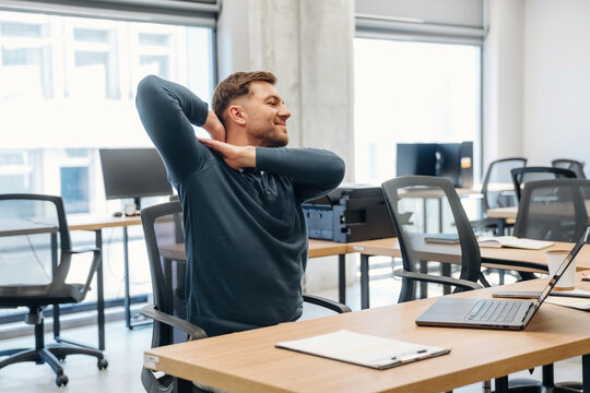 Male office worker in blue sweater stretches in modern workspace with wooden desks, laptop, and empty chairs, bright windows providing natural light