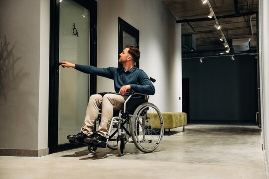 Young man in wheelchair reaching for door handle in modern interior space with concrete flooring and minimalist design elements visible in the background