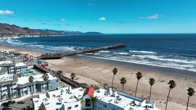 Aerial drone view of Pismo Beach Pier, palm-lined beachfront hotels, and the Pacific coastline on a clear day in Pismo Beach, California, USA.