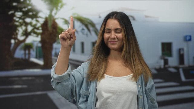 Woman points index finger upward on street crosswalk near white building while wearing white tanktop and denim shirt; confidence direction.