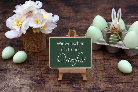 Kleine Tafel mit der Aufschrift 'Wir w&uuml;nschen ein frohes Osterfest' vor einem Eierkarton mit gr&uuml;nen Eiern und einem Osterhasen sowie einem Korb mit wei&szlig;en Blumen auf Holztisch
