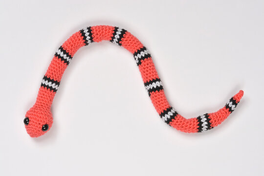 Handmade coral pink amigurumi snake with black and white stripes and small black eyes isolated on a plain white background