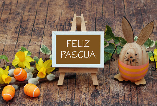 El saludo de Pascua en espa&ntilde;ol &laquo;Feliz Pascua&raquo; sobre un peque&ntilde;o caballete con conejos de madera y narcisos sobre madera r&uacute;stica.