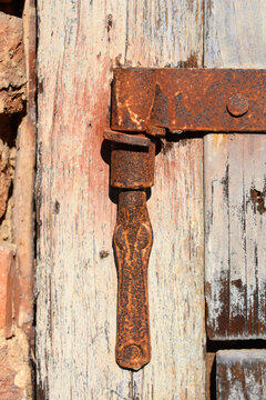 Close up of a very old and rusty metal hinge attached to a weathered wooden door with peeling paint in front of a brick wall