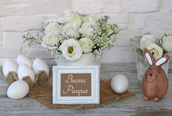 Auguri di Pasqua italiani “Buona Pasqua” in una cornice bianca con rose, uova di Pasqua e coniglietto pasquale davanti a un muro di mattoni. © Racamani
