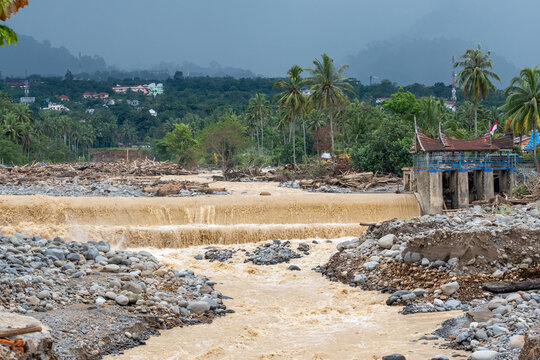 Flooded river with muddy water flowing over small dam in tropical village, showing erosion, debris and environmental impact after heavy rain