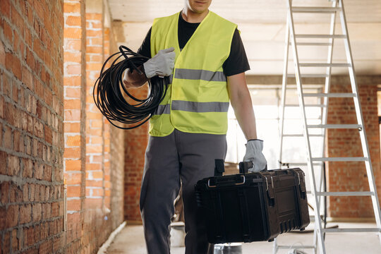 Male construction worker wearing a yellow safety vest carries a toolbox and electrical cables inside a building under renovation with exposed brick walls