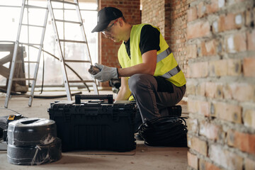 Male construction worker in safety vest examines tools from a black toolbox while sitting on the floor of a building under renovation with a ladder nearby © standret