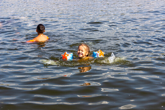 fair-haired girl in swimming armbands swims in river happily squints, laughs, splashes, portrait, muddies water, swims, childhood, rest, teenager, view straight, wet hair, pigtails, summer, day, sunny