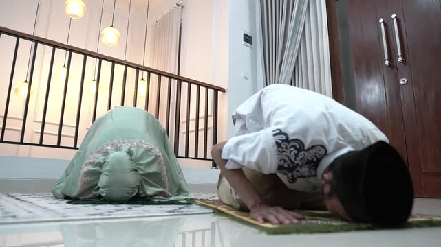Asian couple praying at home, woman in green prayer robe and hijab, man in white shirt and black cap bowing and prostrating on mat