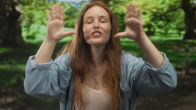 Woman in denim shirt makes heart shape gesture with hands over lips in green park; romance affection joy.