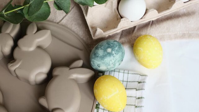 White and colorful Easter eggs with bunny molds and eucalyptus sprig on a table.