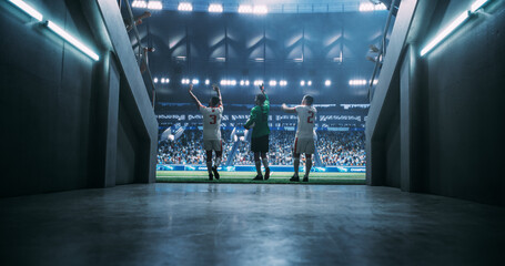 Three Male Soccer Players Exit the Arena Tunnel and Step Toward the Pitch, Saluting Spectators Beneath Floodlights Before Kickoff at a Premier Professional Championship Match Game in a Packed Stadium. © Gorodenkoff