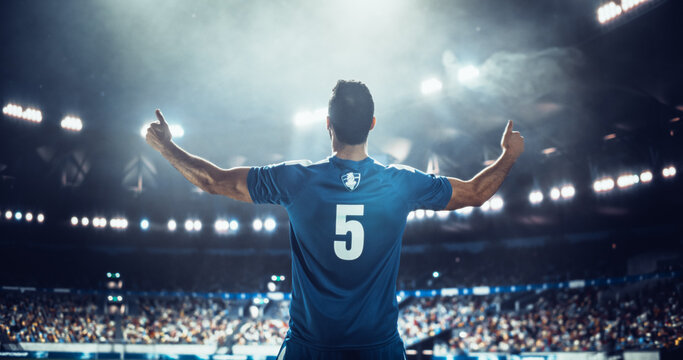 Male Soccer Player Raises Both Arms Toward the Crowd From the Pitch During a Professional International Football Match Game in a Packed World Stadium, Capturing the Scale of a Major Euro Tournament