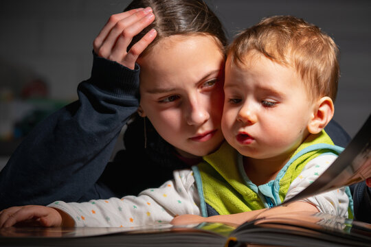 Authentic candid moment of older sister closely supporting her toddler brother while reading, showing deep emotional connection, care, and natural unposed family interaction.
