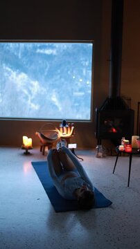 Woman practicing gentle yoga near a fireplace in a cozy mountain house during winter. Warm candlelight, calm atmosphere, slow living lifestyle, evening relaxation indoors