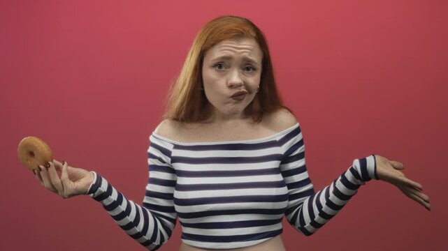 Redhead woman holding a glazed donut, shrugging shoulders while making an uncertain face in studio with pink backdrop and crop top; temptation indecision.