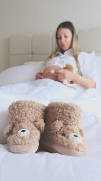 Expectant mother moisturizing her belly with lotion, with cute teddy bear baby shoes in the foreground