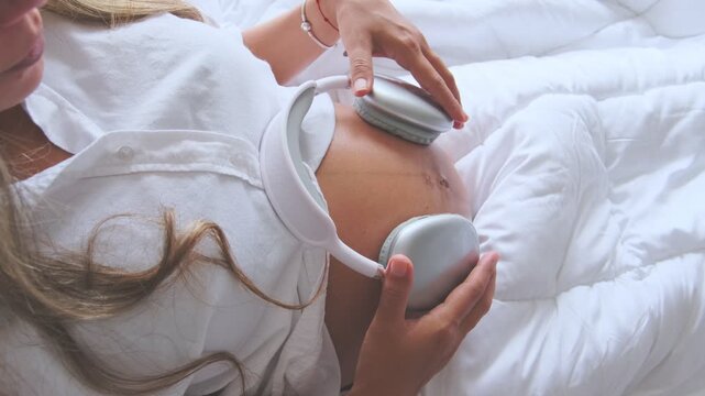 Expectant mother lying in bed holding headphones on her pregnant belly for the unborn baby to listen