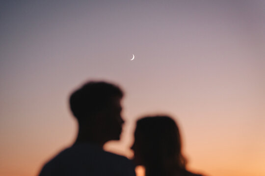 Blurred silhouettes of a couple against a sunset sky with crescent moo