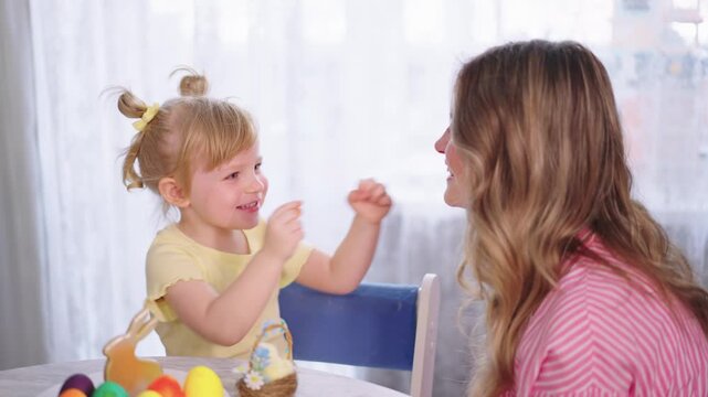 Mother smiles at child during playful kitchen time. Child laughs, claps hands, engages warmly with parent. Soft light fills cozy, lived-in home setting. Perfect for family, parenting