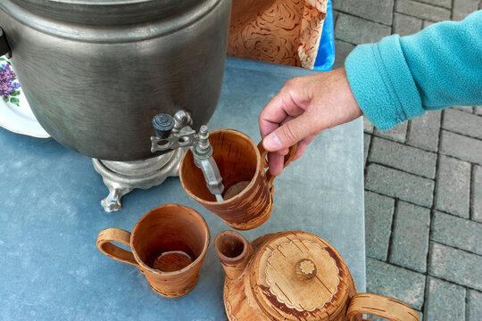 A person is pouring a hot drink from a traditional samovar into a wooden mug.