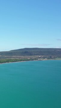 A tranquil view of turquoise waters meeting a lush, green coastline under a clear blue sky in the Isola Varano nature reserve, Italy.